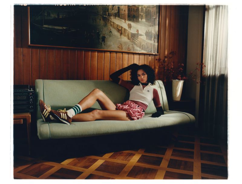 Woman lounges on green couch in retro Adidas athletic wear. An elegant vintage interior with wood paneling and parquet flooring.