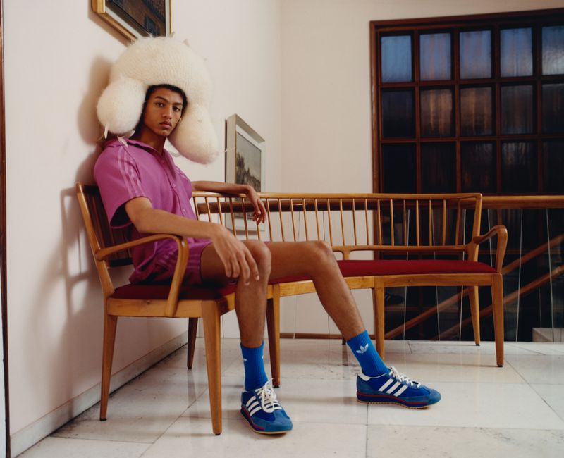Young adult in pink shirt, shorts, blue socks, and sneakers sits on a chair with an oversized, white, fluffy hat indoors.