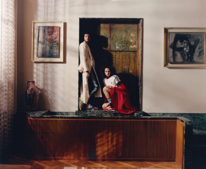 Two figures pose indoors near art and a wooden cabinet. One stands in a white suit, while the other crouches in sports apparel.