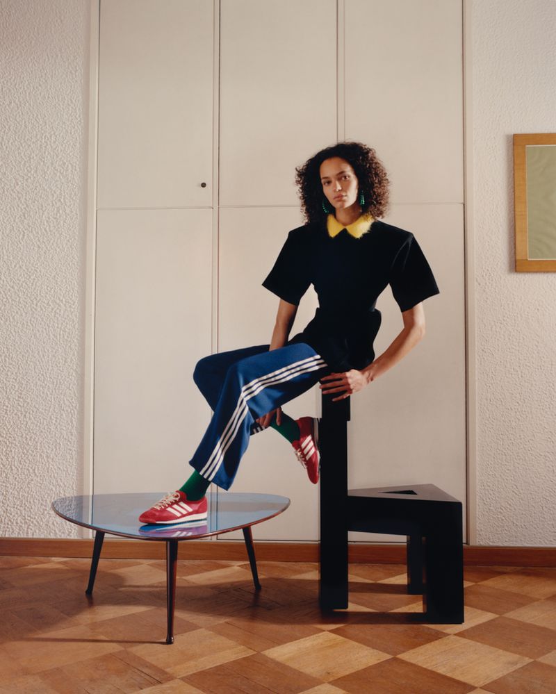 A woman with curly hair poses in a black top and blue Adidas track pants, seated in an indoor setting with a retro aesthetic.