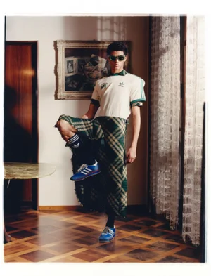 Model posing indoors in a cream retro Adidas jersey, green plaid tartan kilt, dark sunglasses, and blue sneakers on parquet flooring.