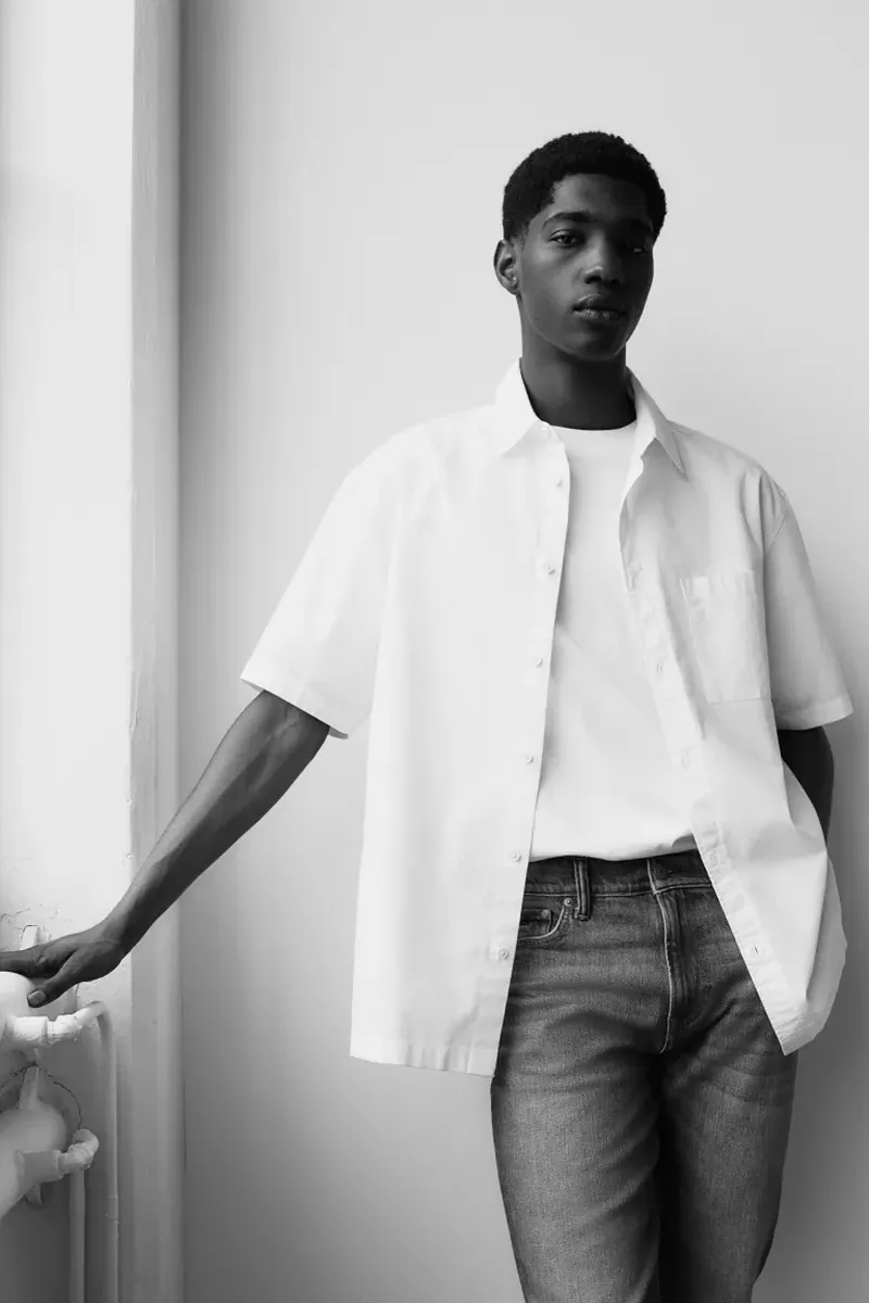 Black male model posing in an open white short-sleeve button-down shirt, white t-shirt, and jeans against a plain wall.