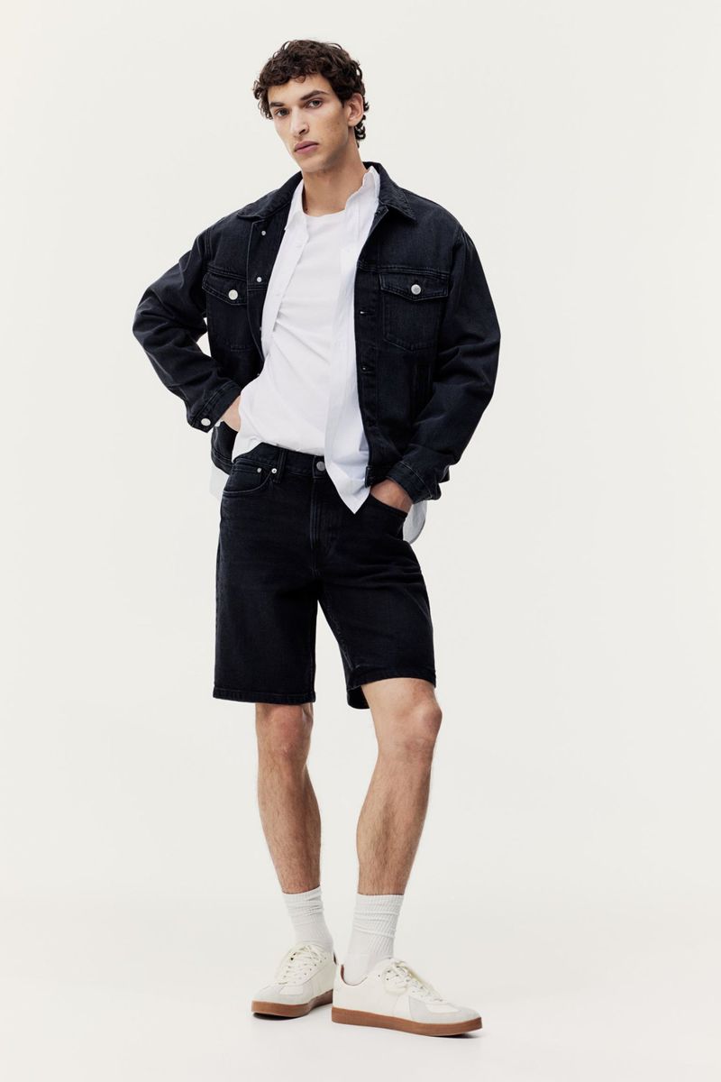 Full-length shot of a brunette man in a dark denim jacket, white shirt, dark denim shorts, white socks, and sneakers, in a studio.
