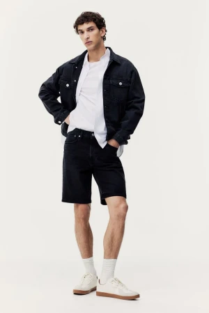Full-length shot of a brunette man in a dark denim jacket, white shirt, dark denim shorts, white socks, and sneakers, in a studio.