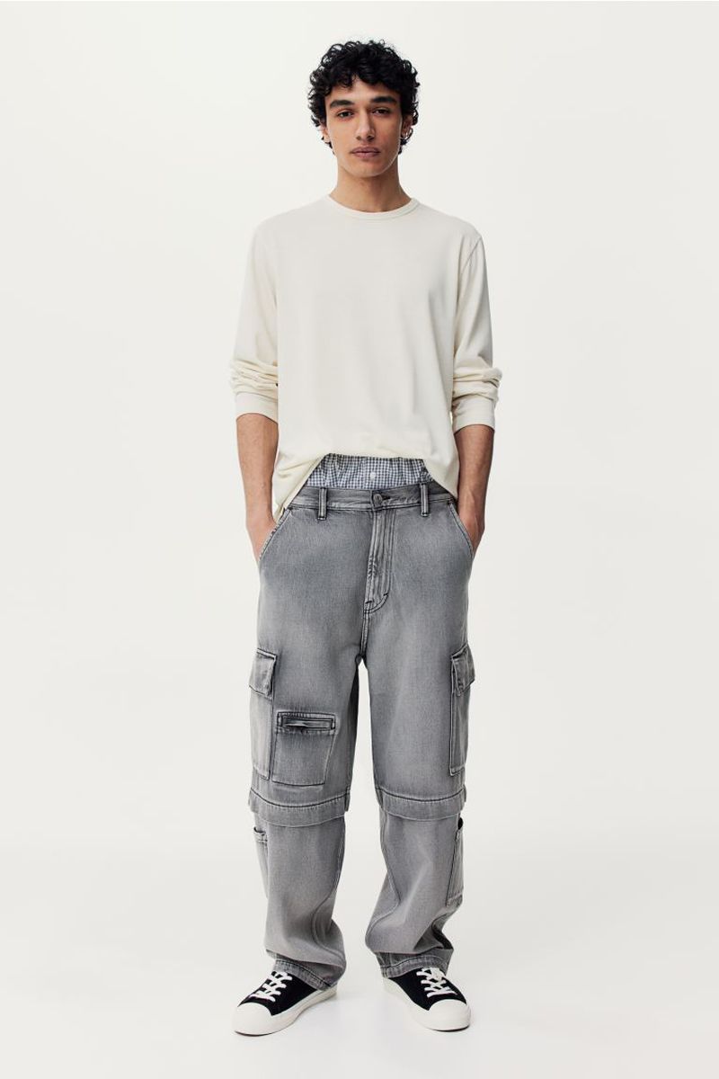 Man in a long-sleeved shirt and gray cargo pants, standing in front of a white backdrop, modeling casual fashion and streetwear with black sneakers.