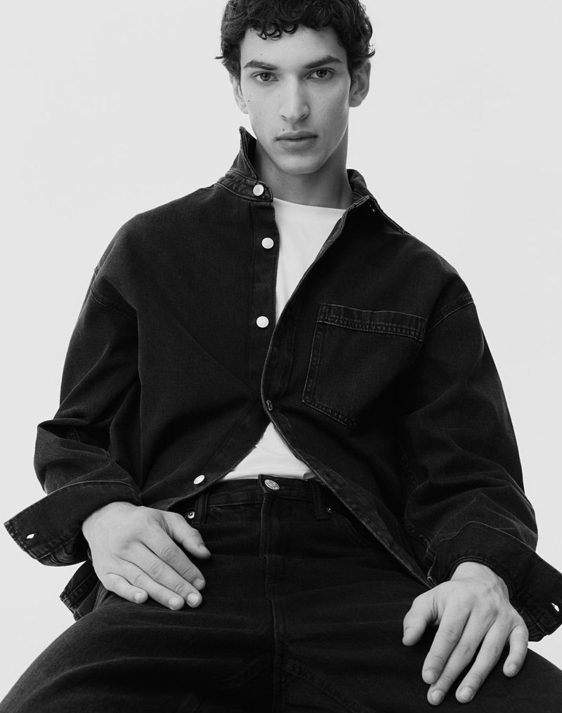 Model seated in an oversized black denim button-up shirt and black jeans against a plain white studio background.