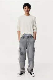 Full-length portrait of a male model wearing grey cargo jeans and a cream long-sleeve t-shirt against a plain white background.