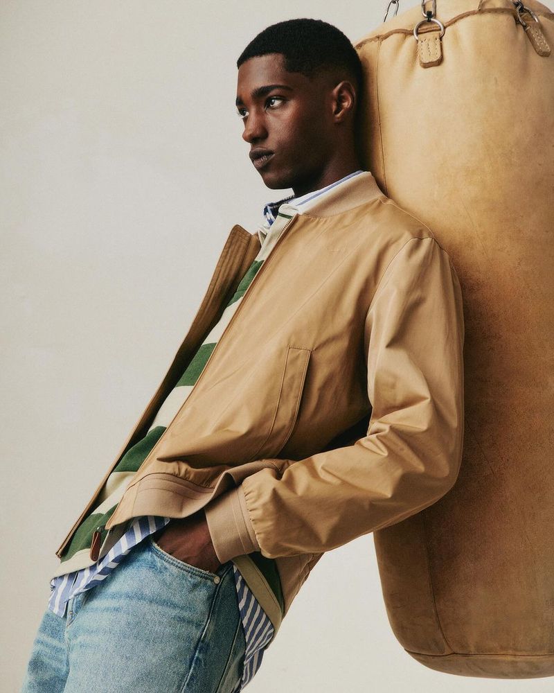 Tan jacket and blue jeans on a dark-skinned man leaning against a tan punching bag in a studio setting.