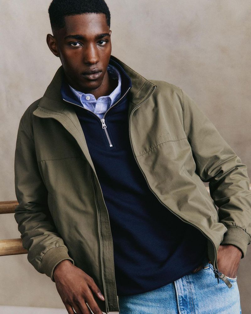 A dark-skinned male model in a light-green jacket, navy polo shirt, and light-blue jeans strikes a casual pose against a neutral backdrop.