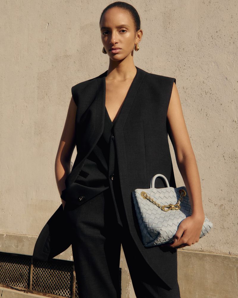 Fashionable woman in a black vest and pants holds a light blue woven tote bag, posing against a neutral wall in natural light.