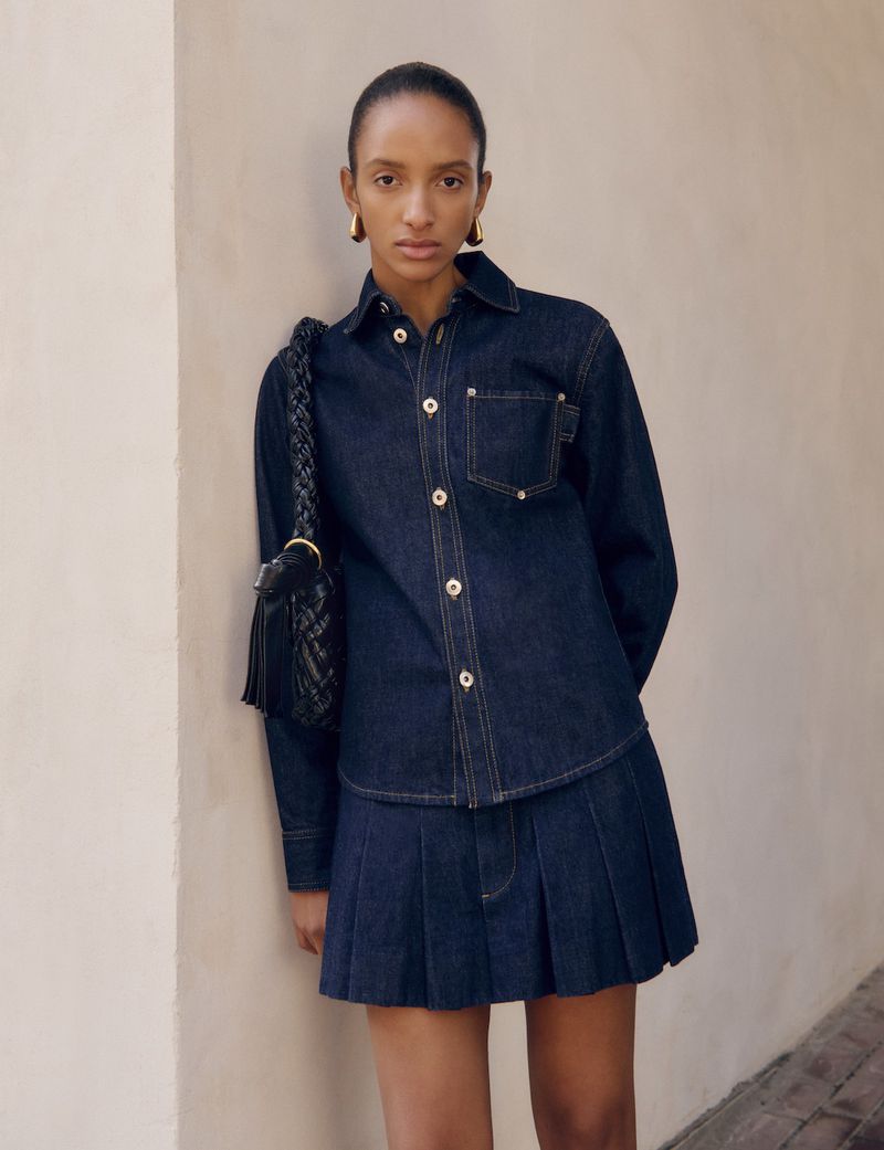 Fashion shot of a woman in a dark denim collared shirt and pleated skirt with a woven shoulder bag leaning against a wall.