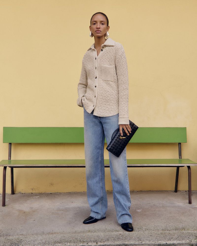 A stylish woman in a knit shirt and jeans holds a clutch bag in front of a yellow wall and green bench, showcasing fashion.
