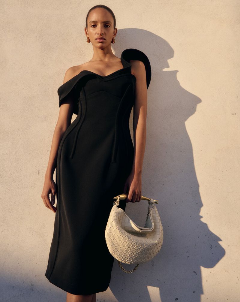 Elegant woman in a black off-the-shoulder midi dress holding a woven white purse with a gold handle in a studio setting.