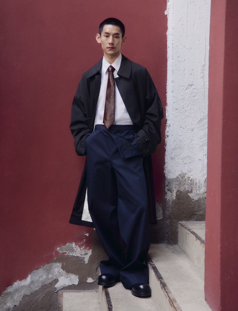 Asian man in an overcoat, dress shirt, necktie, and wide-legged pants standing on stairs against a red and white wall indoors.