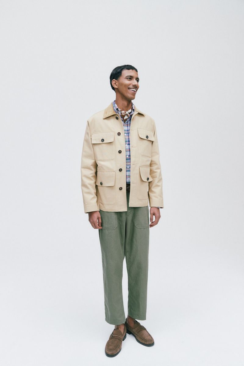 A full shot of a smiling man wearing a tan jacket, plaid shirt, and olive pants, standing against a white background in a studio setting.