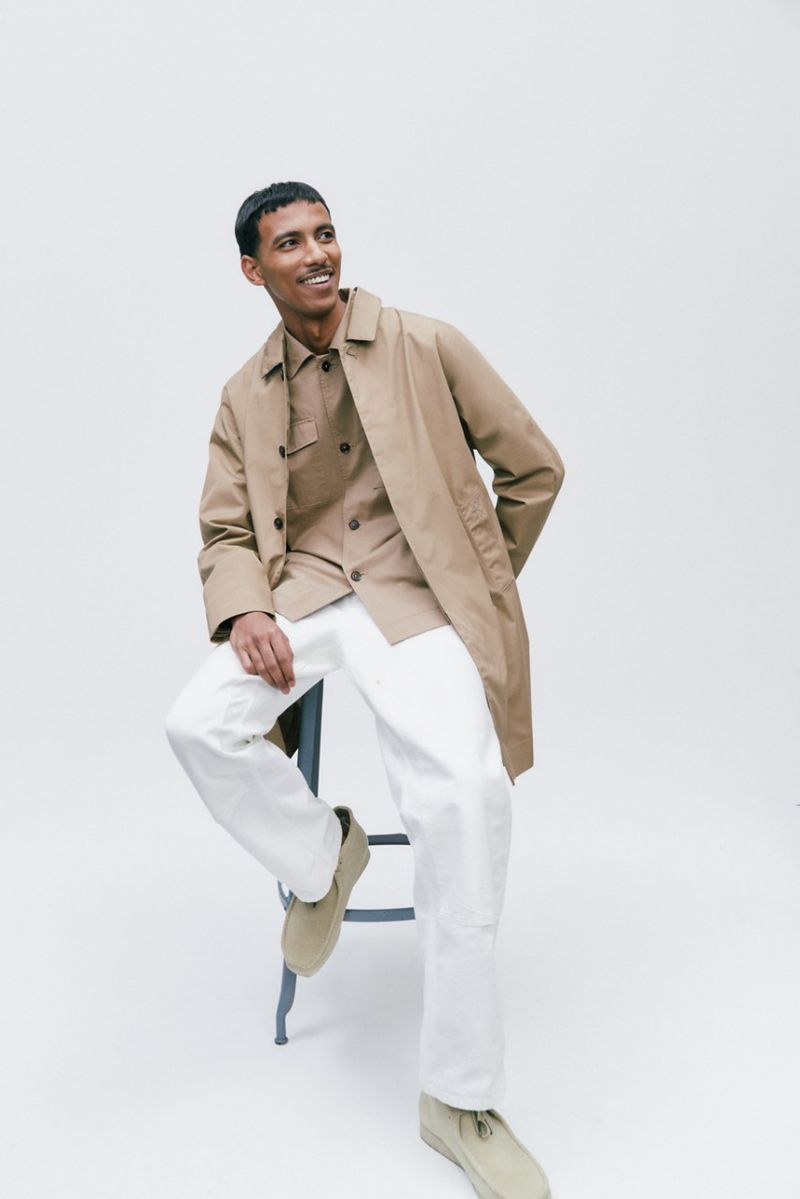 Man in a tan trench coat sitting on a stool in a white studio, smiling and looking up and to the left with tan shoes.