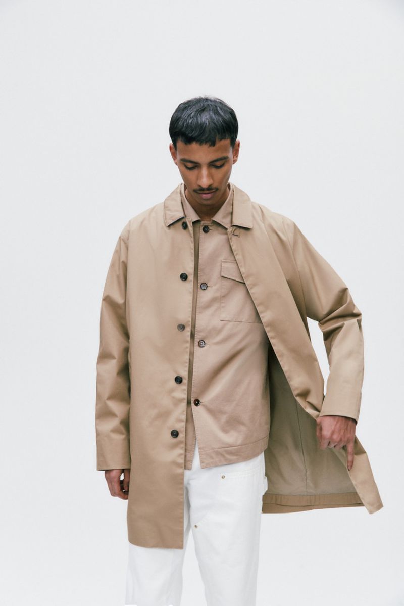 Man wearing a khaki trench coat and white jeans poses against a white backdrop in a fashion setting. The style is minimalist.