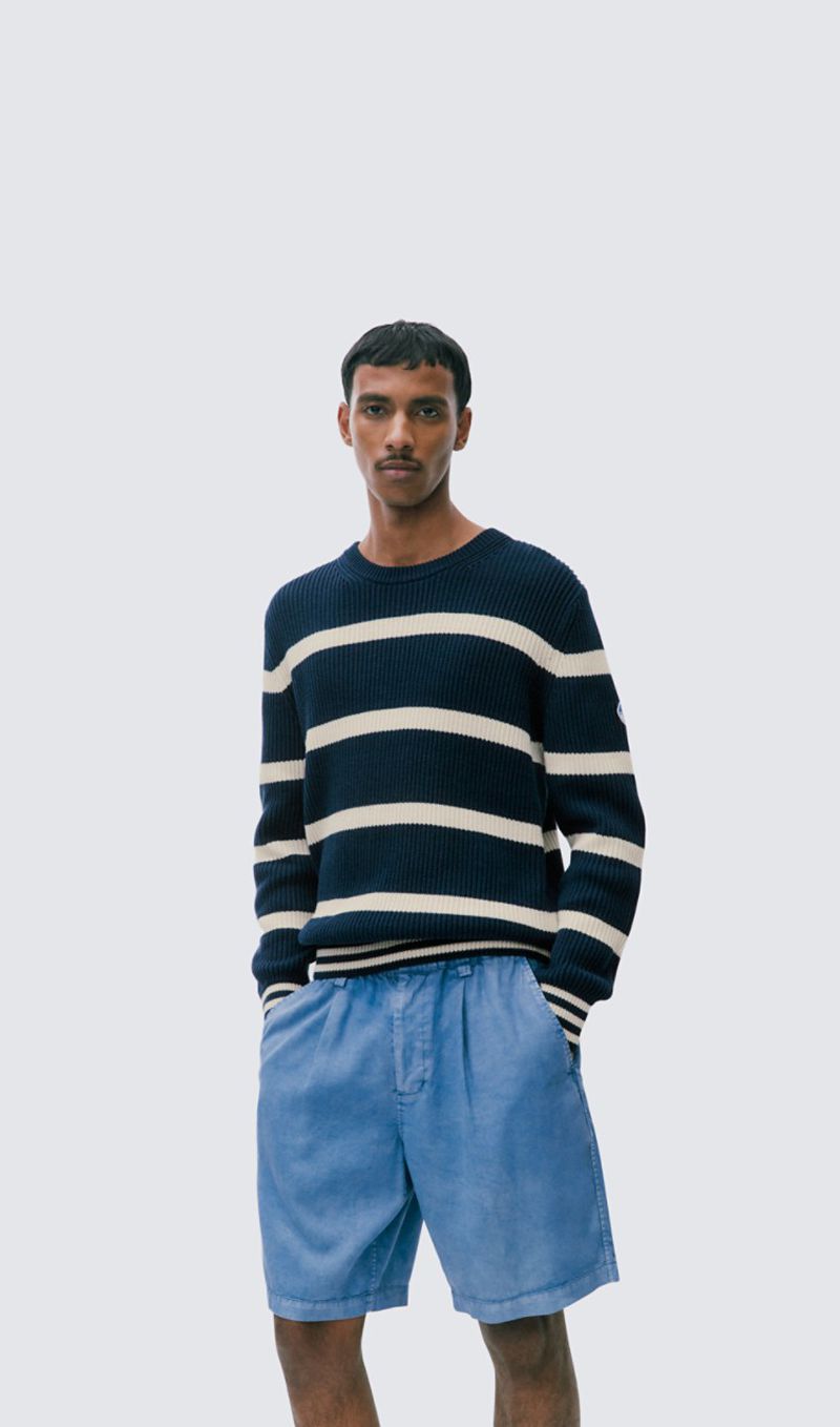 Studio shot of a man wearing a navy and white striped sweater and blue shorts. He stands against a gradient blue and white background.