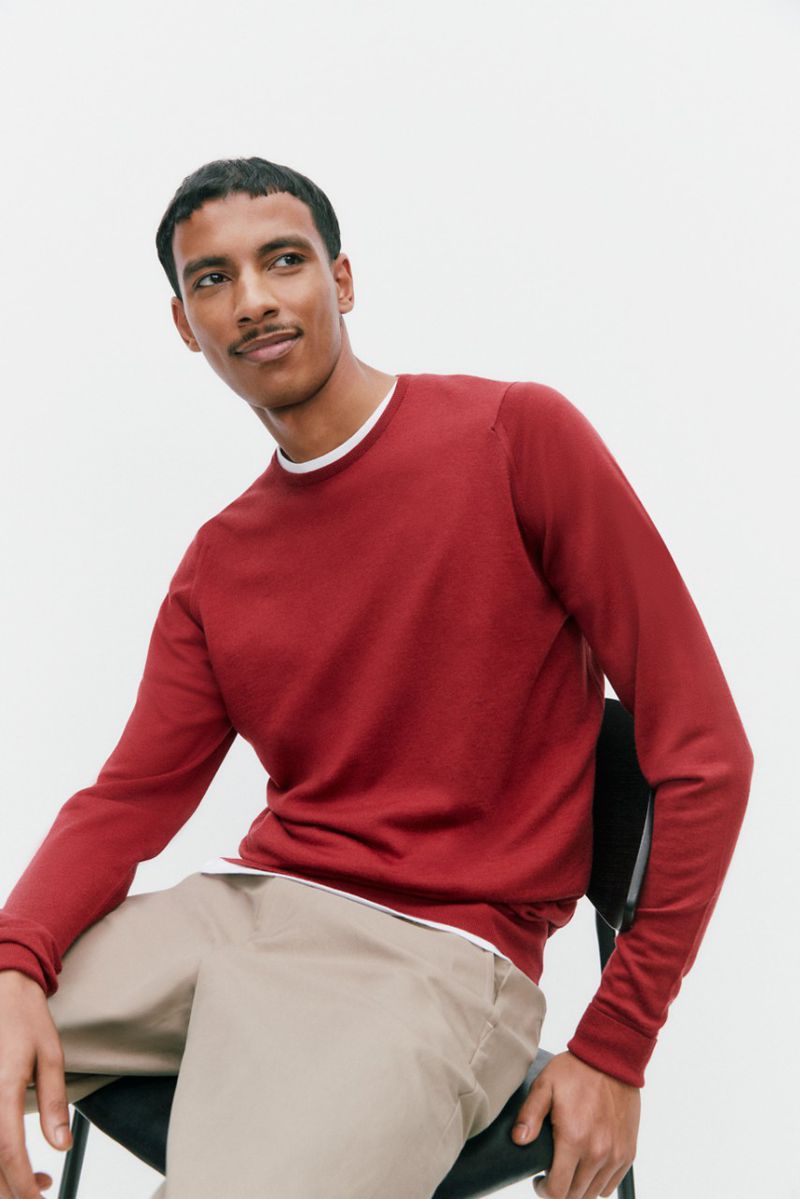 Model Mohamed Hassan seated in a black chair wearing a red crewneck sweater and beige trousers against a white seamless background.