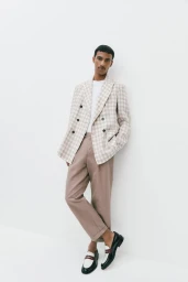 Model Mohamed Hassan wearing a checkered double-breasted blazer and taupe trousers against a bright white studio backdrop.