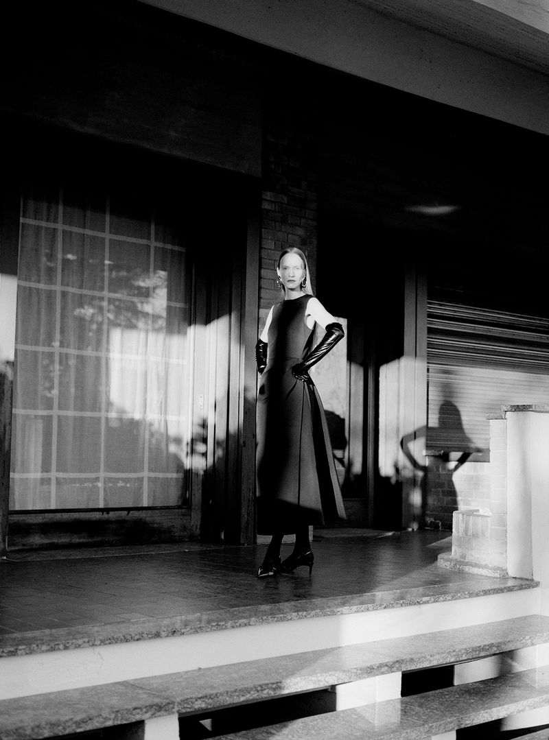 Model standing on a porch wearing a dark dress and long gloves, photographed by Davide Sometti for Maison Cogo FW2425.
