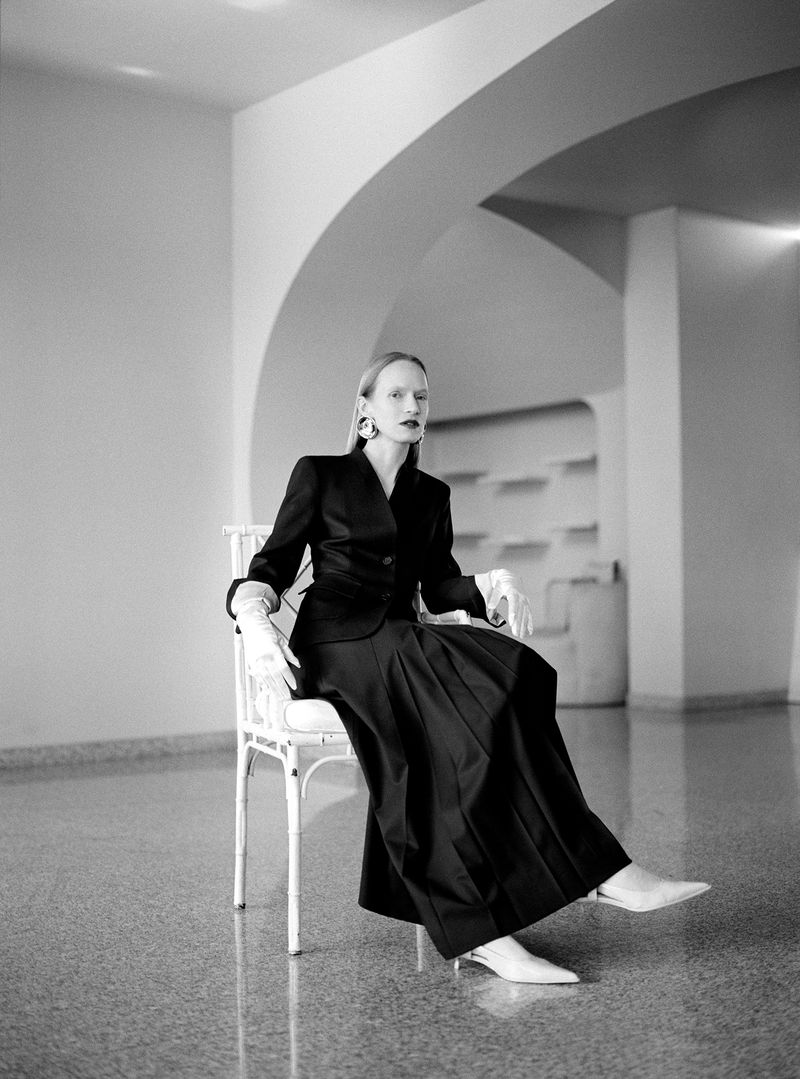 Elegant woman in black blazer and skirt seated on a white chair indoors, creating a sophisticated fashion editorial in black and white.