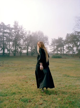 Woman in a black outfit stands in a grassy field looking back at the camera, trees and fog in the background, contemplative mood.