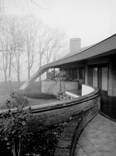 Black and white image showing modern architecture of a brick house with a curved wall, a covered outdoor area, and trees in the background.