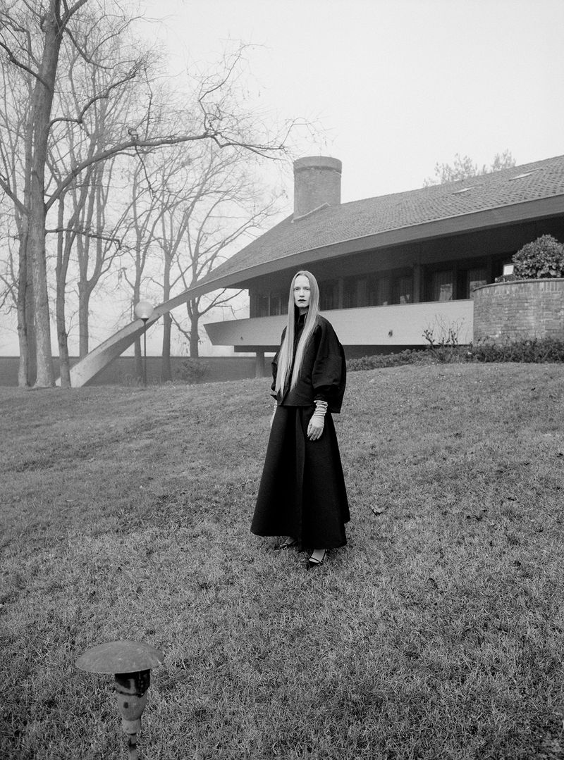 Elegant adult female in a black outfit stands on a lawn near a modern house with bare trees and overcast sky, creating a striking fashion editorial.