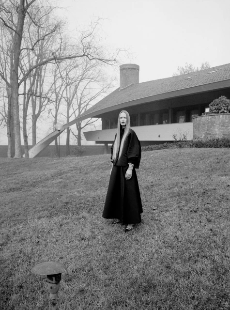 Elegant adult female in a black outfit stands on a lawn near a modern house with bare trees and overcast sky, creating a striking fashion editorial.