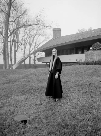 Elegant adult female in a black outfit stands on a lawn near a modern house with bare trees and overcast sky, creating a striking fashion editorial.