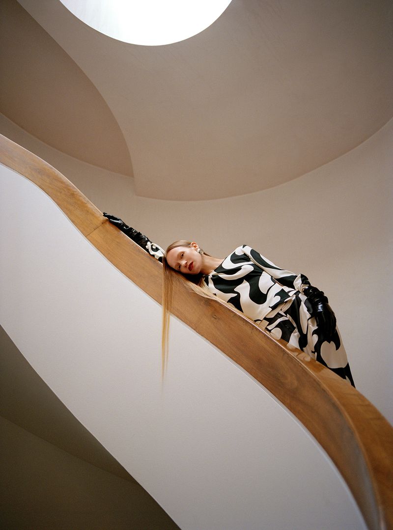 Young woman in a black and white dress reclining on a wooden staircase with white walls, illuminated by natural light from above.