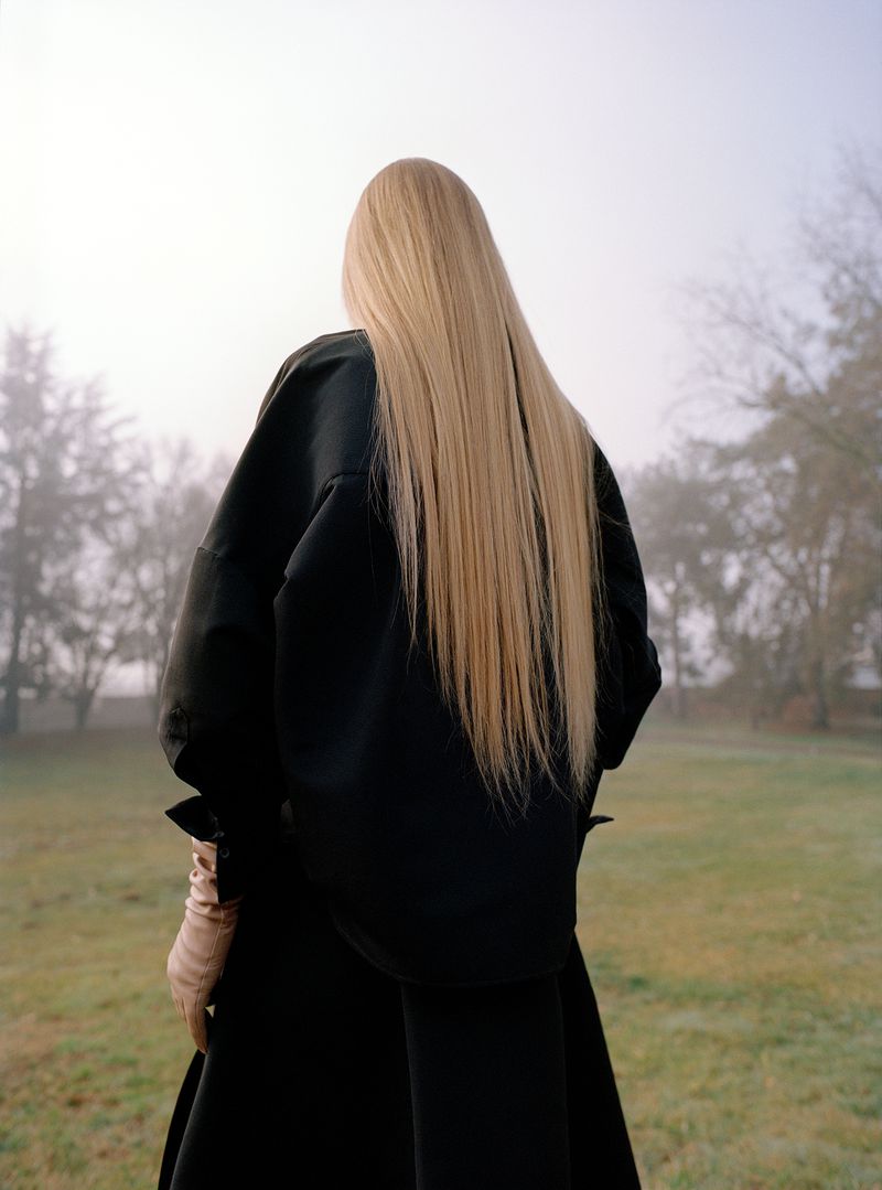 Woman with long blonde hair stands in a field wearing a black coat and tan gloves, facing away from the camera on a foggy day.