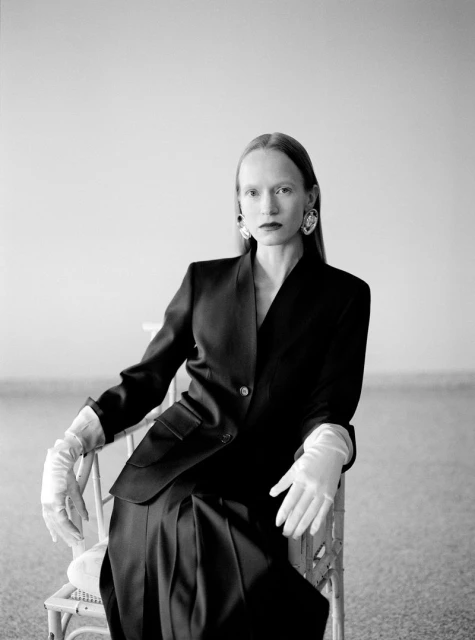 Elegant woman in a black suit and white gloves sits on a chair in a monochrome portrait, fashionably styled with large earrings.