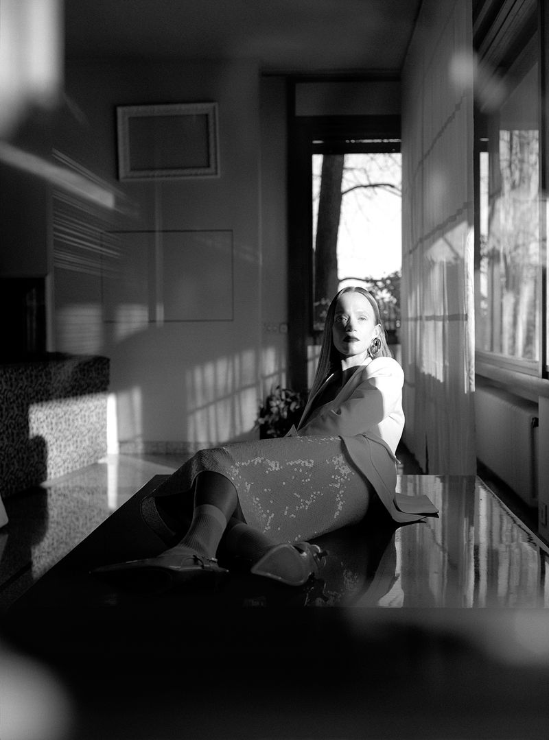 A woman with long hair sits on a table in a room with a window and white walls, a painting, and a decorative plant, in black and white.