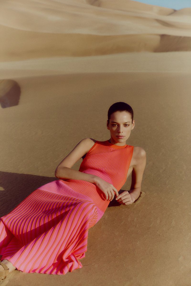Woman in pink-and-orange sleeveless dress laying on sand dunes in an outdoor fashion shot with eye-level framing and natural lighting.