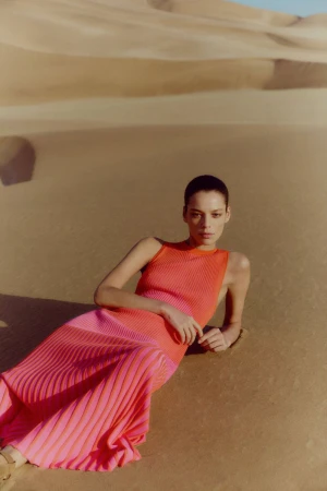 Woman in pink-and-orange sleeveless dress laying on sand dunes in an outdoor fashion shot with eye-level framing and natural lighting.