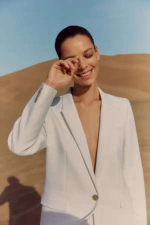 Woman in a white blazer smiles while shielding her eyes in a desert landscape under a clear blue sky, fashion photography.