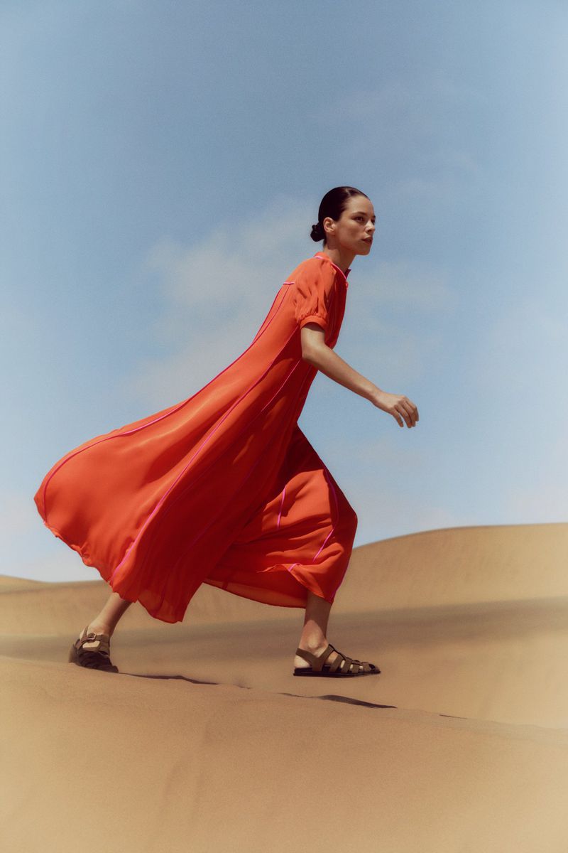 Model Nigina Sharipova walks across sand dunes wearing a billowing bright orange maxi dress and flat sandals under a clear sky.