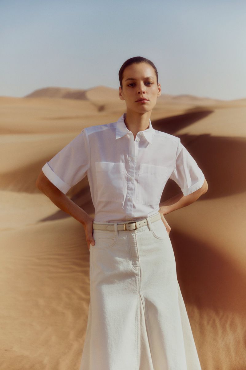 Woman in white shirt and skirt posing in a desert landscape. Fashion shot with sun and sand dunes, creating a bright, natural scene.