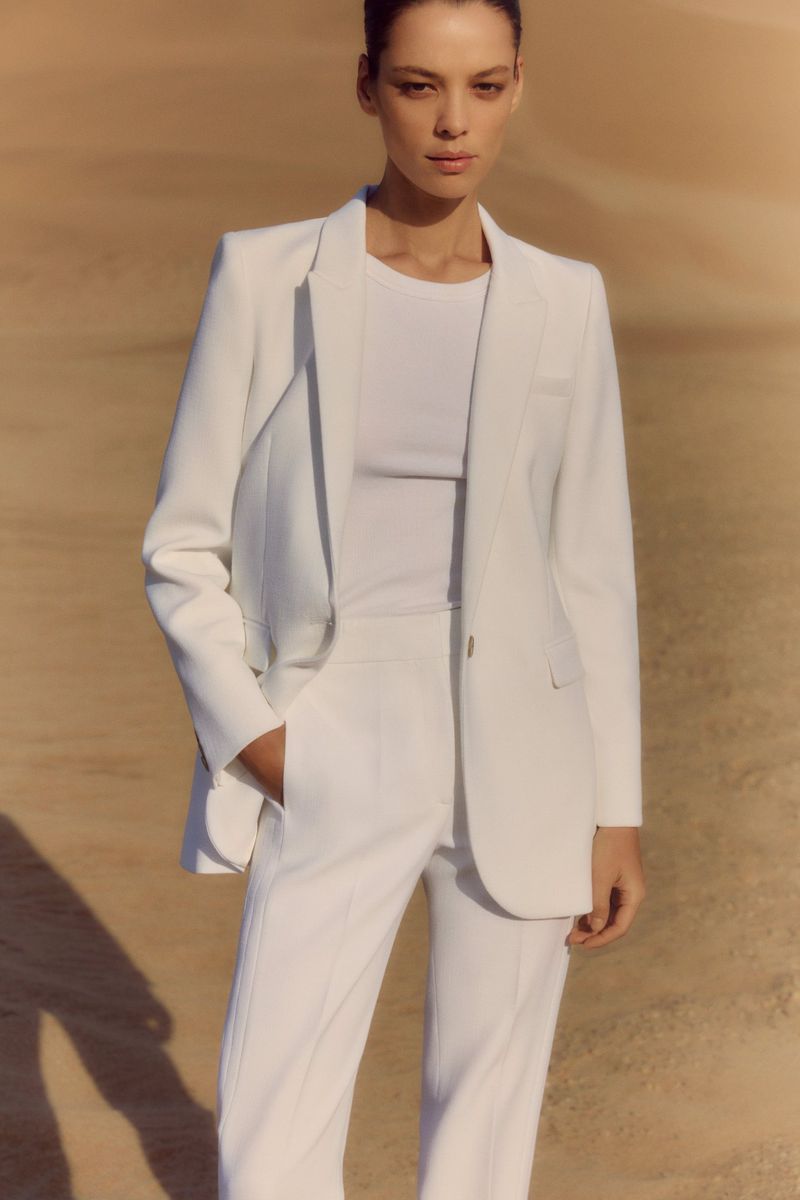 Fashion shot of a woman in a white blazer and pants, standing outdoors against a neutral backdrop, showcasing modern, minimalist style.