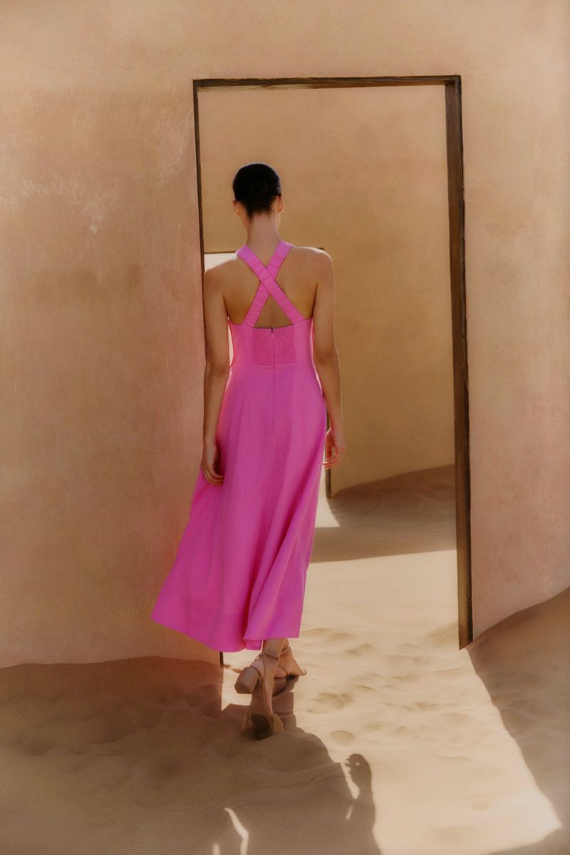 Back view of a woman in a bright pink sleeveless dress walking through a doorway in a hallway with warm beige walls and floor.