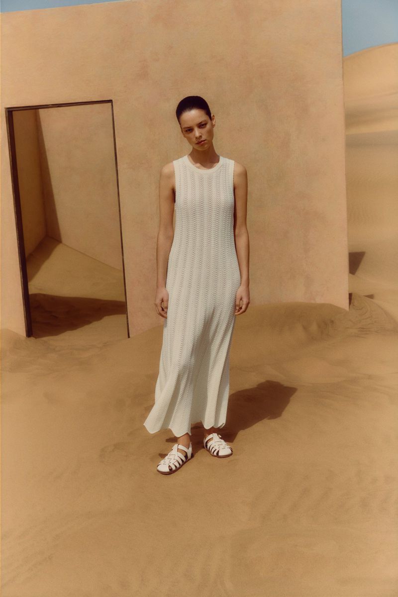 A young adult woman stands in a full-length white dress with neutral-toned sandals on a sand dune surface in front of a doorway in a tan wall.