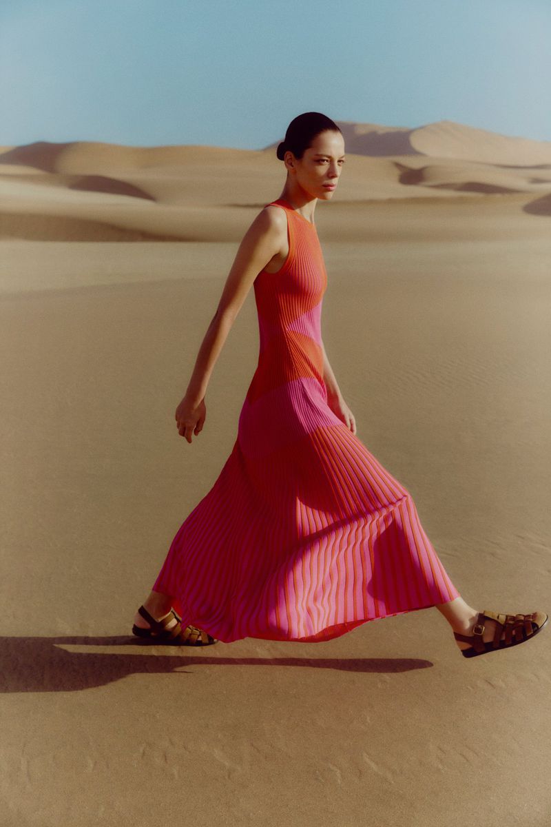 An adult female model walks through a sandy desert landscape wearing a colorful, sleeveless dress and dark sandals against a blue sky.