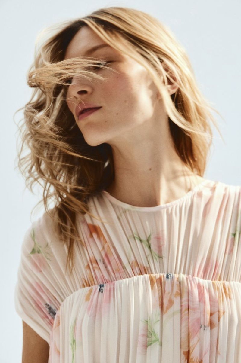 Close-up of a blonde woman in a floral-patterned dress against a blue sky, with windblown hair creating a soft, airy aesthetic.