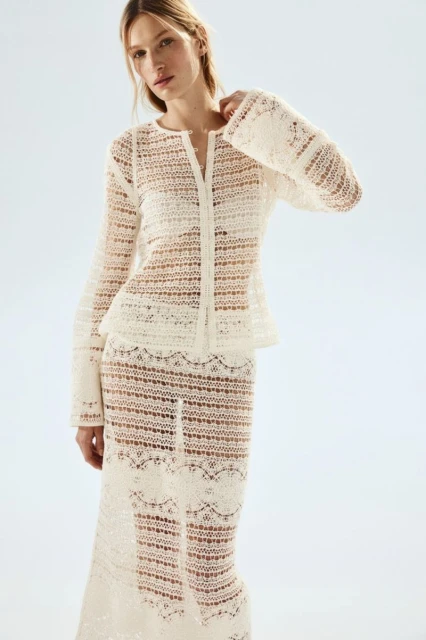 Adult female fashion model poses in a semi-sheer, crocheted long-sleeved top and maxi skirt against a neutral backdrop in studio lighting.