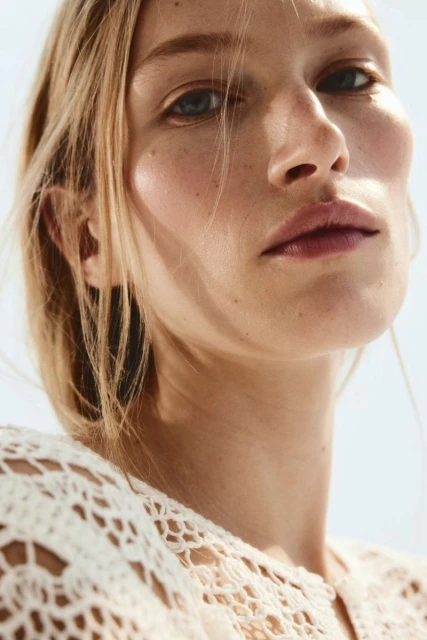 Close-up portrait of a blonde adult woman with fair skin, wearing a white, patterned, crocheted top, against a light-blue background.