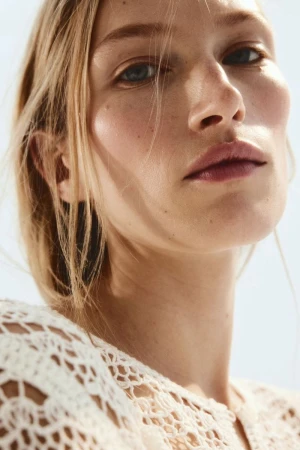 Close-up portrait of a blonde adult woman with fair skin, wearing a white, patterned, crocheted top, against a light-blue background.