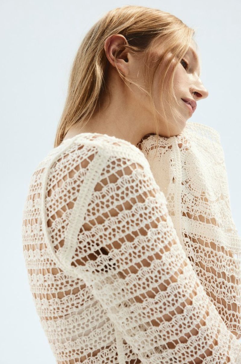 Close-up studio shot of a woman wearing a cream-colored, long-sleeved, crocheted shirt in a minimalist fashion setting under bright lighting.