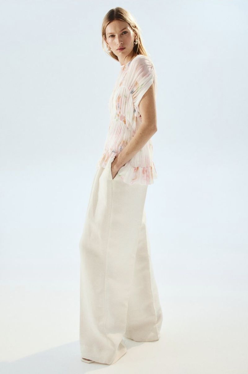 Model Liz Kennedy wearing wide-leg cream trousers and a pleated floral sleeveless blouse against a clean white backdrop.